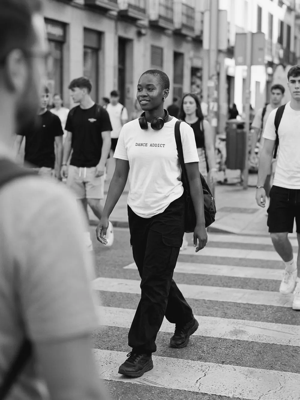Chica cruzando un paso de cebra con la camiseta blanca DANCE ADDICT y los cascos al cuello, en blanco y negro