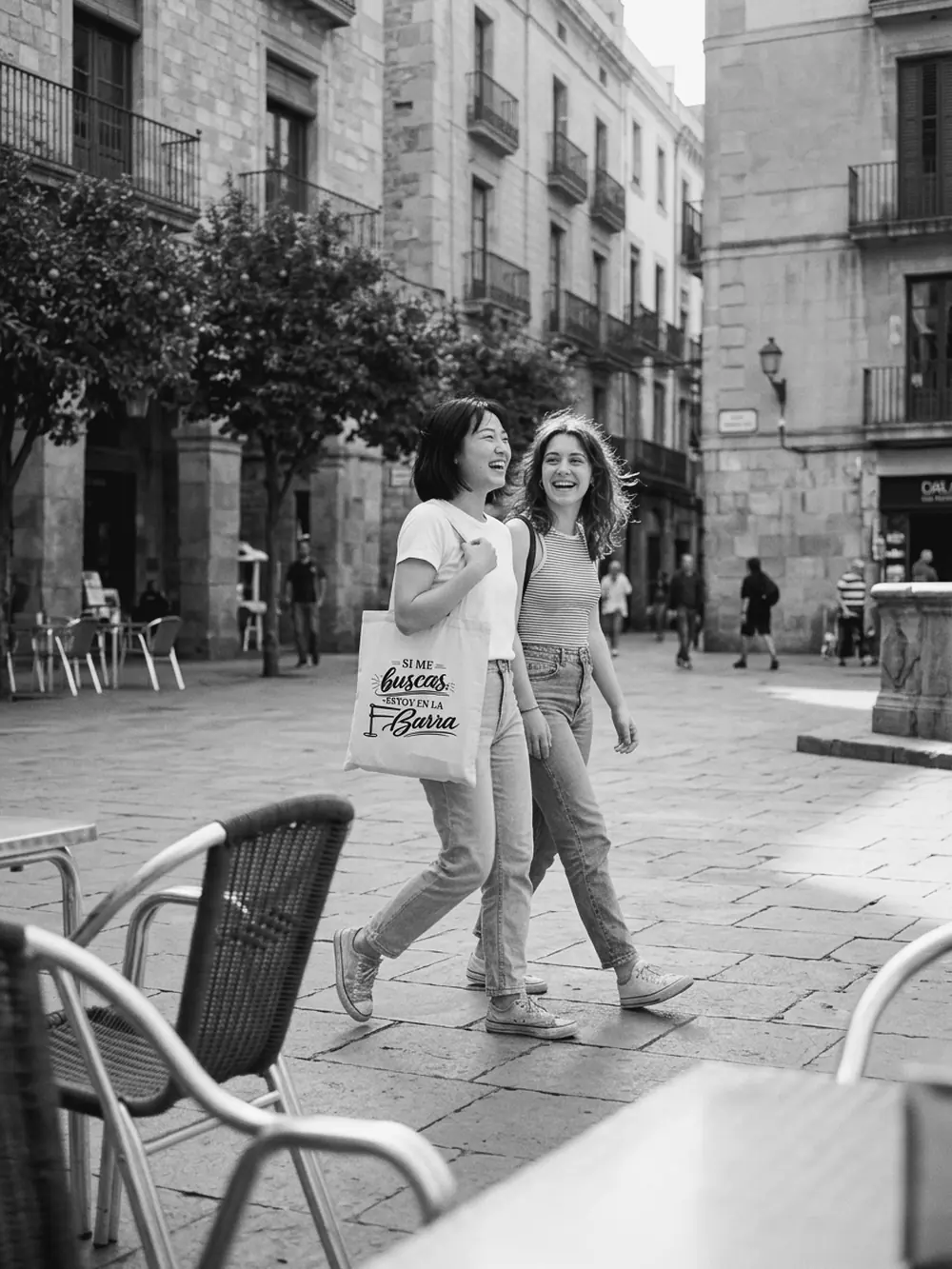 Dos amigas riendo por una plaza del casco antiguo, una con la tote bag Si me buscas, estoy en la barra al hombro